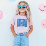 A young girl with blonde hair wearing round sunglasses, a light pink graphic t-shirt that says 'FOOTBALL sister' in blue, and light blue jeans, standing against a white wall.