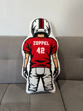 A custom-shaped throw pillow shaped like a football player from behind, wearing a red jersey with 'ZOPPEL 42' on the back, resting on a gray fabric sofa.