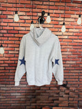 A light heather gray pullover hoodie with two blue star patches on the sleeves, hanging on a hanger against a red brick wall with hanging light bulbs.
