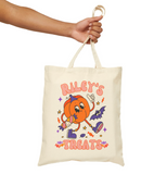 An off-white canvas tote bag with a cartoon pumpkin character and the text 'RILEY'S TREATS' in orange, held by a person's hand against a plain white background.