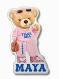 A pillow in the shape of a cartoon teddy bear wearing sunglasses, a pink sweatsuit with "TYLER HILL" and it's zip code printed on it, and pink sneakers. The bear is holding a drink and a brown purse. The name "maya" is printed in blue at the bottom.