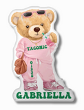 A pillow in the shape of a cartoon teddy bear wearing sunglasses, a pink sweatsuit with "TACONIC" and it's zip code printed on it, and pink sneakers. The bear is holding a drink and a brown purse. The name "gabriella" is printed in green at the bottom.