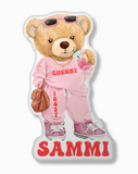 A pillow in the shape of a cartoon teddy bear wearing sunglasses, a pink sweatsuit with "CHENNY" and "18427" printed on it, and pink sneakers. The bear is holding a drink and a brown purse. The name "SAMMI" is printed in red at the bottom.