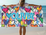 A colorful beach towel with dripping graffiti heart design in multi colors. The name "OLIVIA" is prominently displayed in light blue block letters.
