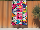 A colorful beach towel with dripping graffiti heart design in multi colors. The name "BELLA" is prominently displayed in pink block letters.