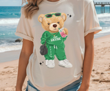 Tan t-shirt featuring a cartoon teddy bear wearing sunglasses and a green tracksuit with the text "Camp Taconic" on it's shirt, and the camp's zip code down the pants leg. The bear is holding a pink soft drink and a black hand bag, and wearing white sneakers.