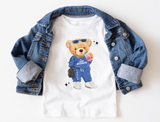 white t-shirt featuring a cartoon teddy bear wearing sunglasses and a blue tracksuit with the text "Camp Westmont" on it's shirt, and the camp's zip code down the pants leg. The bear is holding a pink soft drink and a black hand bag, and wearing white sneakers.