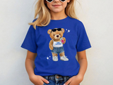 Blue t-shirt featuring a cartoon teddy bear wearing sunglasses a white tank top, and denim shorts, with the text "Camp Starlight" on it's shirt. The bear is holding a pink soft drink and a black hand bag, and wearing white sneakers.