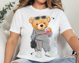 White t-shirt featuring a cartoon teddy bear wearing sunglasses and a grey tracksuit with the text "Timber Tops" on it's shirt, and the camp's zip code down the pants leg. The bear is holding a pink soft drink and a black hand bag, and wearing white sneakers.