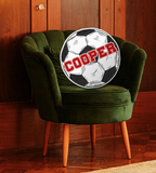 Soccer ball pillow with the name 'COOPER' printed across it in red, sitting on a green chair. 