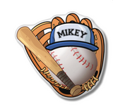 Baseball glove pillow holding a baseball wearing a  blue hat that has a name, and a baseball bat laying across it. 
