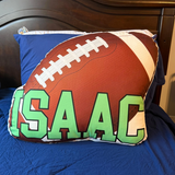 Football-shaped pillow with 'ISAAC' printed on it, placed on a bed.