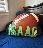 Football-shaped pillow with 'ISAAC' printed on it, placed on a bed.