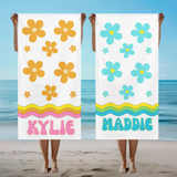 Two beach towels with daisy patterns and names 'Kylie' and 'Maddie' held up on a beach. Kylie towel has orange daisies and Maddie's towel has turquoise daisies. 