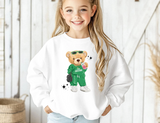 A young girl with long blonde hair smiling while wearing a white crewneck sweatshirt featuring a cartoon graphic of a teddy bear in a green outfit holding a drink. The background is a soft-focus white wooden wall.