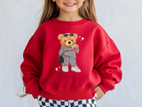 A young girl wearing a bright red crewneck sweatshirt with a cartoon teddy bear graphic holding a drink. The sweatshirt features a camp name across the chest and it's zip code down the bears leg. 