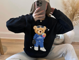 A person taking a mirror selfie wearing a black sweatshirt with a large teddy bear graphic on the front, paired with light gray sweatpants in a home setting.