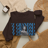 A dark brown sweatshirt laid flat on a light beige surface, featuring a graphic print with the word 'ZZ GRANDMA' repeated in blue and a black-and-white photo of a child. The sweatshirt is styled with a cream knit blanket, a woven straw bag, a tan knit bag, and a small candle jar.