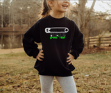 A young girl wearing a black crewneck sweatshirt with a white safety pin graphic and green 'Green Lane' text, standing outdoors in a park setting.