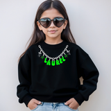 A young girl with long dark hair and heart-shaped sunglasses wears a black crewneck sweatshirt featuring a bright green 'LAUREL' text design with silver chain and star graphics on the chest, paired with light blue jeans against a plain light gray background.