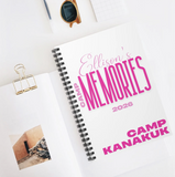 A spiral-bound notebook laid open on a white surface, printed with the text 'Ellison's MEMORIES 2026' and 'CAMP KANAKUK' in pink lettering. The scene includes a gold binder clip, sunglasses, and a small photo insert as styled props.