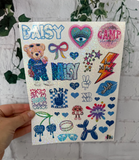 A hand holds a sheet of glittery stickers featuring various designs like a teddy bear, hearts, stars, and text such as 'DAISY' and 'CAMP' against a white brick wall with green ivy.
