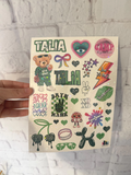 A hand holds a sheet of glittery stickers featuring various designs including the word 'TALIA' in green, a teddy bear, a heart with an eye, a lightning bolt, and other colorful doodles, set against a white brick wall background.