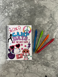 A spiral-bound notebook with a colorful 'Camp Days Are The Best Days!' cover design lies on a gray marble surface next to four colored pens in blue, green, orange, and red.