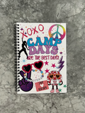 A spiral-bound notebook with a white cover decorated with colorful retro 'Camp Days' graphics, including peace signs, stars, sunglasses, and cartoon characters, photographed on a gray marble surface.