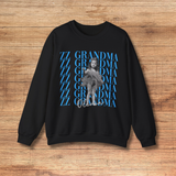 A black crewneck sweatshirt featuring a graphic print of a grayscale photo of a girl in a tutu over a repeating blue 'ZZ GRANDMA' text with 'Olivia' written in white script below. The sweatshirt is laid flat on a light brown wooden background.