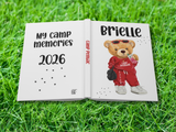 A custom hardcover photo book lying open on green grass, featuring a white cover with the text 'My camp memories 2026' on the left page and a cartoon teddy bear wearing a red tracksuit with the name 'Brielle' on the right page.