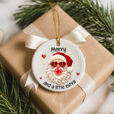 A round white Christmas ornament with a printed Santa face, red hat, and the phrase “Merry and a little extra,” hanging from a gold string on top of a brown gift box with a cream ribbon, surrounded by pine branches.