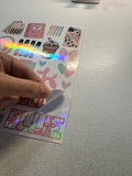 A hand peeling a sticker from a sheet of decorative stickers on a light gray textured desk. The stickers feature pink and multicolor designs including hearts, bows, a coffee cup, and patterned accessories, with one sticker showing a holographic rainbow sheen.