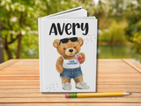 A personalized hardcover notebook with a white cover featuring a cartoon teddy bear wearing sunglasses, denim shorts, and a white tank top that says 'camp starlight'. The name 'Avery' is printed at the top in a black handwritten-style font